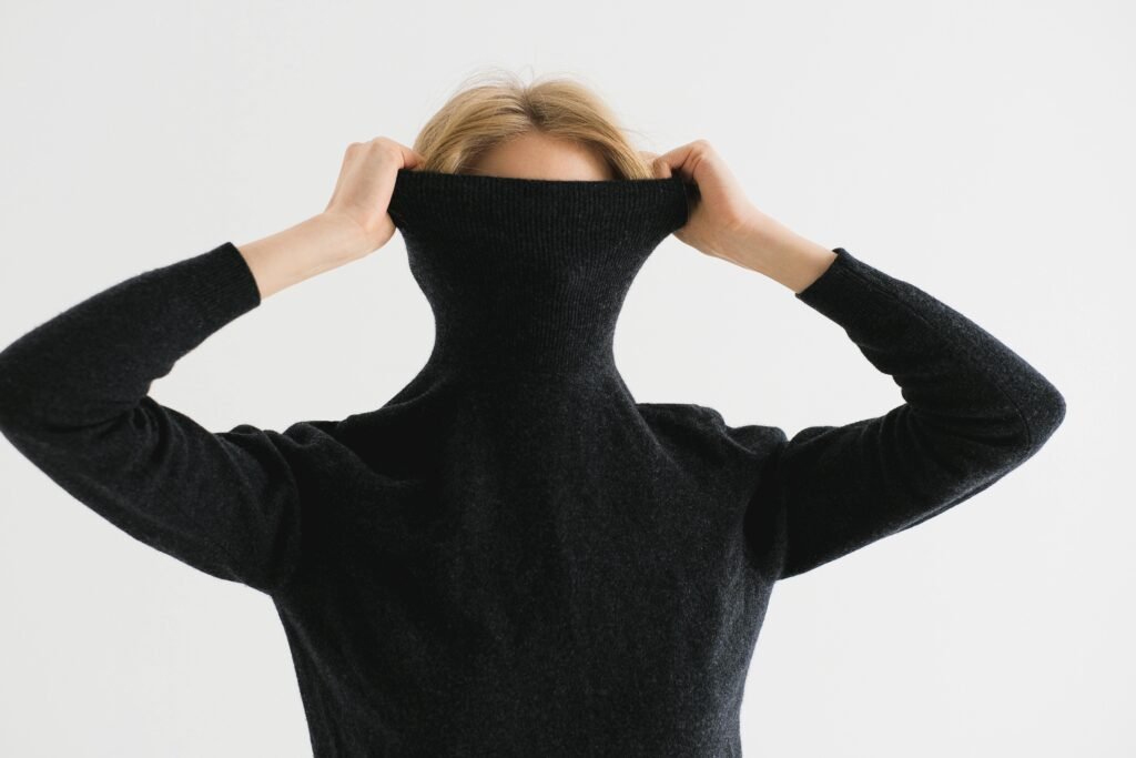A woman playfully hides her face in a black turtleneck sweater, expressing a shy demeanor.